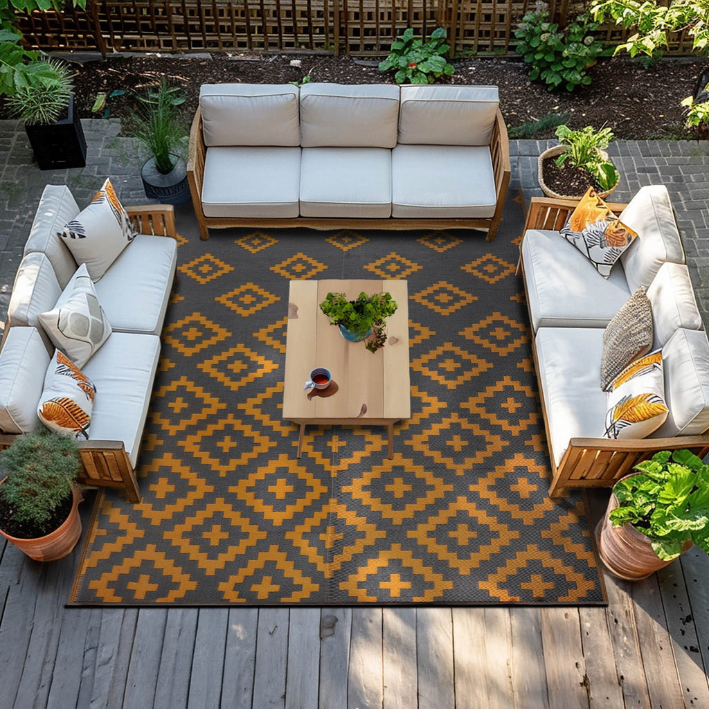 Green Decore Bettina Outdoor Rug in Charcoal Grey and Yellow Ochre displayed on a wooden patio, surrounded by light beige cushioned wooden sofas and tropical-themed cushions, with a wooden coffee table and lush greenery in the background.