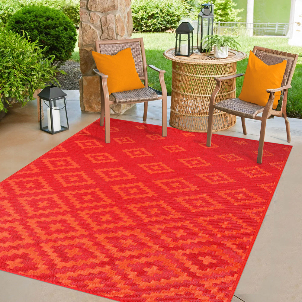 Green Decore Bettina Red and Orange Outdoor Rug featuring a bold geometric pattern, placed on a patio with wicker chairs, orange cushions, a round wicker table, black lanterns, and lush greenery in the background—ideal for garden or balcony décor. 