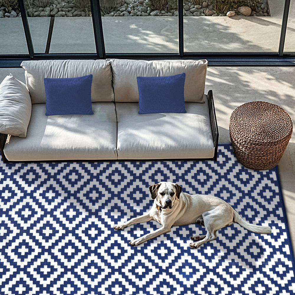 Green Decore Bliss Outdoor Recycled Plastic Rug in Navy Blue and White with geometric diamond pattern, placed in a modern patio setting with a beige sofa, blue cushions, a woven round ottoman, and a dog lounging comfortably on the rug.