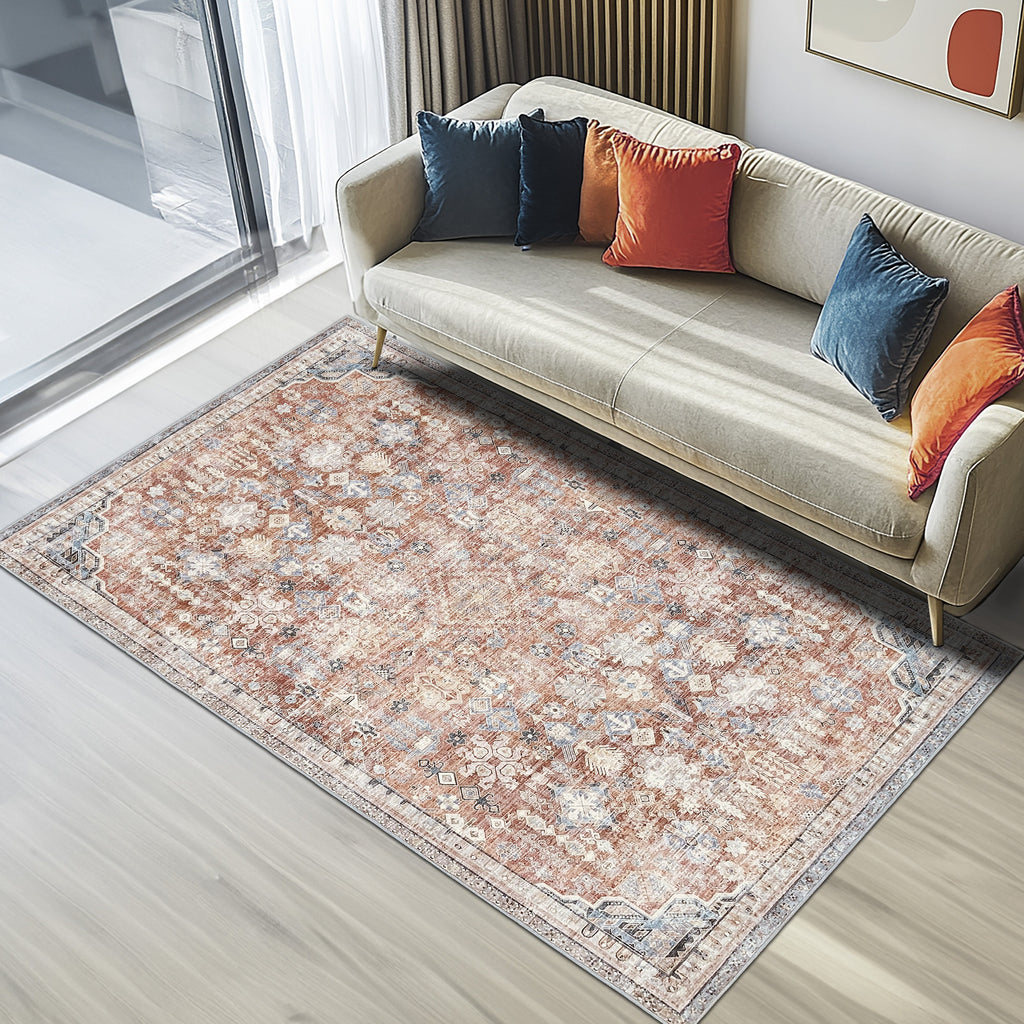 Modern living room with a beige sofa decorated with blue and orange cushions, featuring a vintage-style Green Decore’s vernal Egbert machine washable rug in rust, yellow, and blue tones on light wood flooring beside large windows with sheer curtains