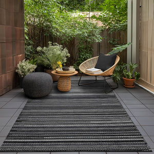 Black Outdoor Rug with striped pattern placed on a tiled patio, styled with a wicker chair, round tables, and potted plants for a chic and serene outdoor seating area.
