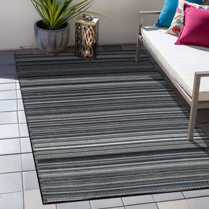 Black Outdoor Rug with subtle striped design, placed on a tiled balcony beside a white bench with colourful cushions and a potted plant, perfect for stylish outdoor lounging.

